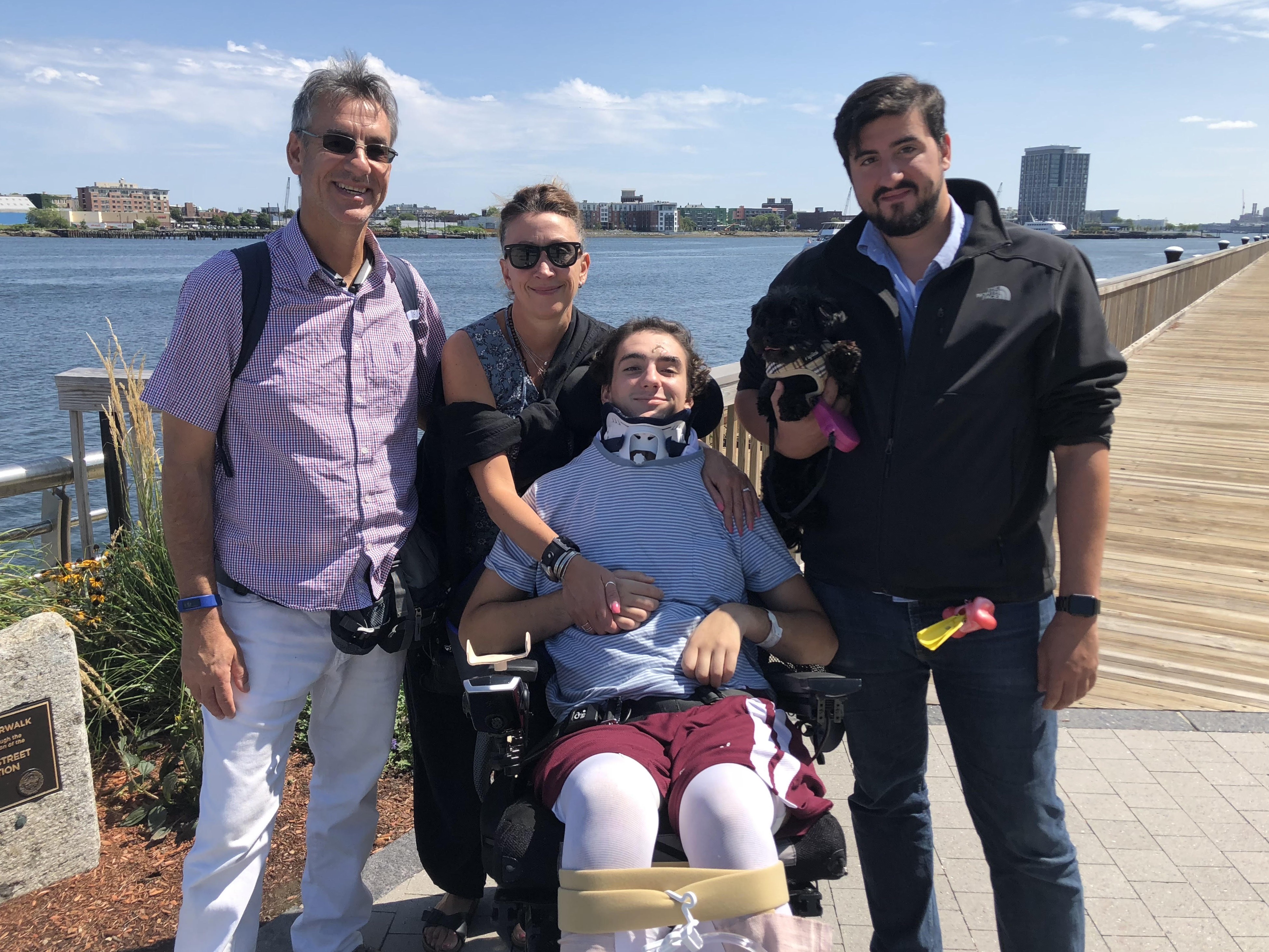 A father, mother, and older brother stand beside a young man in a wheelchair on a boardwalk.