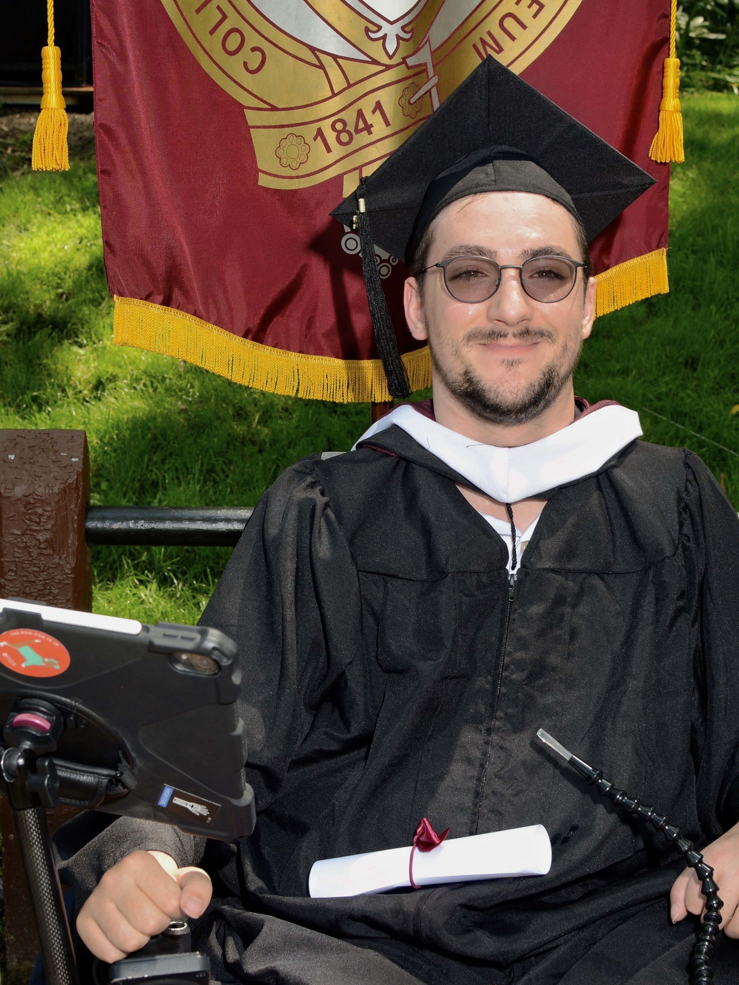 A man wearing sunglasses and a black graduation cap and gown smiles.
