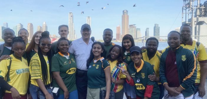 A group of students wearing green and maroon shirts smile in front of a city skyline and flying birds in the sky.