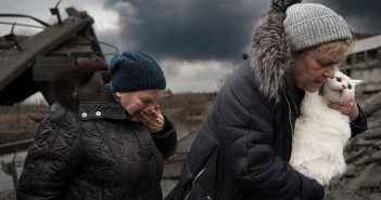 Two women, one holding a white cat in her arms, the other covering her mouth with her hand, flee a battle scene in Irpin, Ukraine