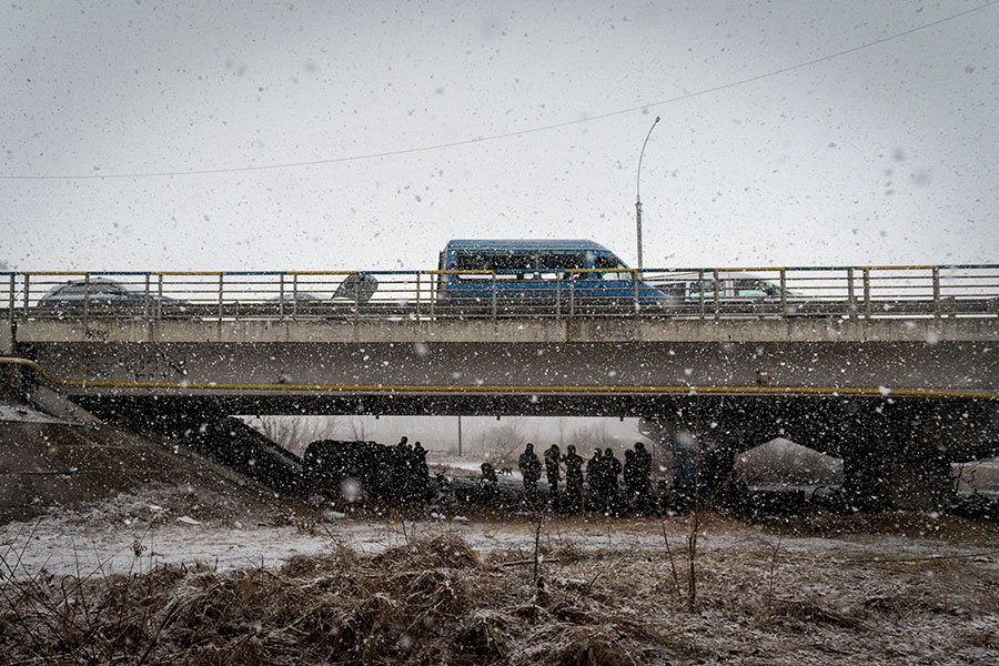Vehicles abandoned atop a damaged bridge as people huddle underneath the bridge and a light snow falls