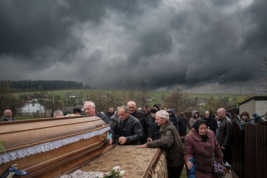 A group of mourners stand outside near a coffin, dark gray storm clouds in the background