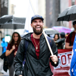 Man smiling while marching in parade, he has a beard and a baseball cap on, people holding umbrellas behind him.