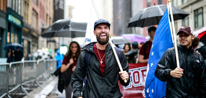 Man smiling while marching in parade, he has a beard and a baseball cap on, people holding umbrellas behind him.