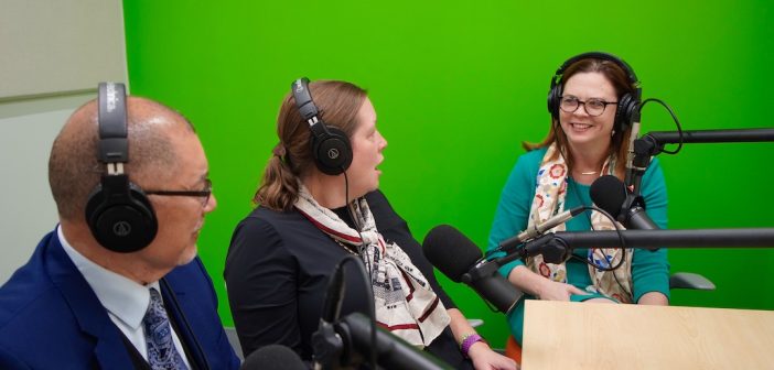 Three seated people wearing headphones speak into microphones.