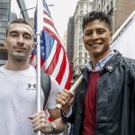 Two men smiling looking at camera. One is holding an American flag in a white shirt, the other is holding another flag that we can not make out wearing a maroon shirt and a black leather jacket.