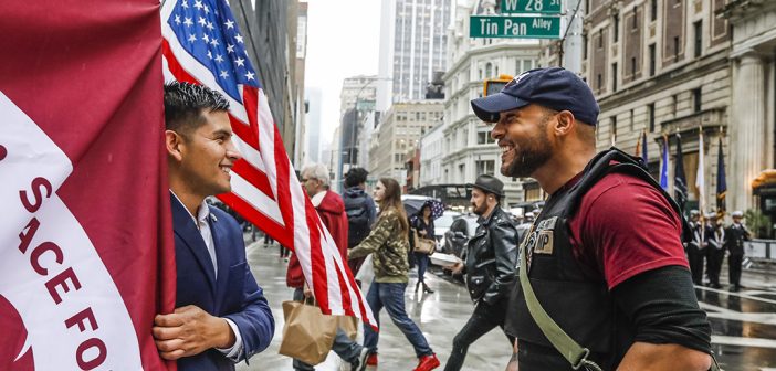 Man in suit holding maroon flag and man in dark blue baseball cap smiling as they speak to each other. There is an American flag in the background and pedestrians.