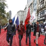 Six men, marching in parade holding different flags. They are walking on a red carpet that fills up the Manhattan street. They are smiling.