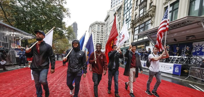 Six men, marching in parade holding different flags. They are walking on a red carpet that fills up the Manhattan street. They are smiling.