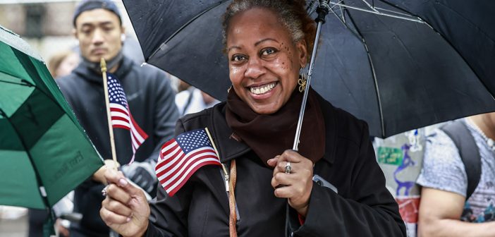 Woman, holding a black umbrella wearing a black coat and a brown scarf. She has a small American flag in her hand and another one stuck on her purse strap.