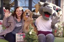 A woman, a golden retriever, and a person dressed in a ram outfit sit on a sofa and smile.