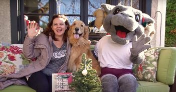 A woman, a golden retriever, and a person dressed in a ram outfit sit on a sofa and smile.