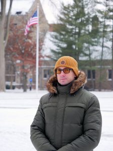 A man with a green jacket and yellowhat stands outside with snow on the ground. An American flag flies on a pole bhind him.