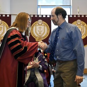 A woman wearing maroon graduation robes shakes the head of a man wearing a format outfit.