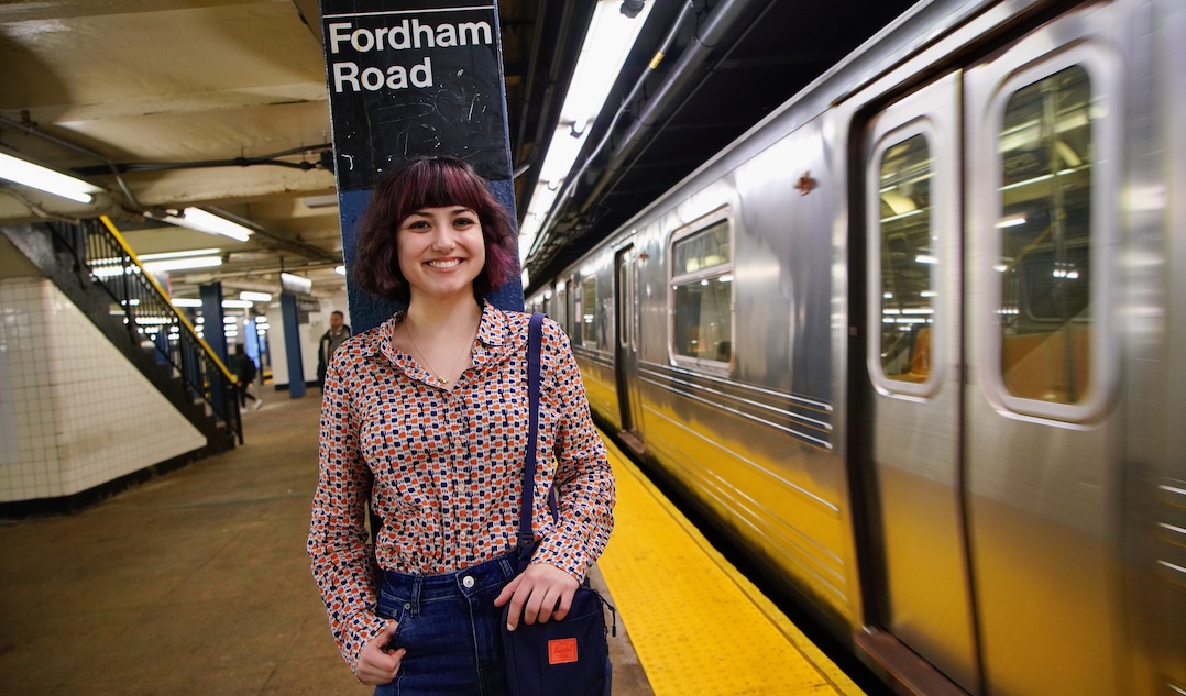 A girl smiles at the camera while a subway train rushes past her on the right.