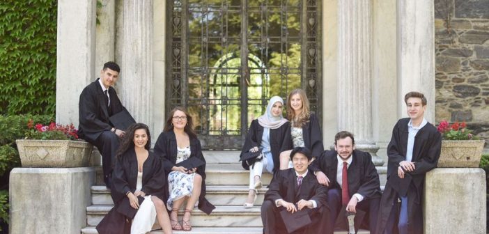 A group of Fordham graduates in cap and gown pose on the steps of Cunniffe House, with its white columns and galss doors