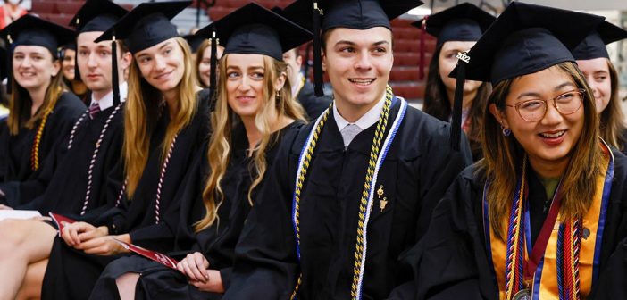 A group of Fordham graduates, in cap and gown, sitting in a line and looking off camera