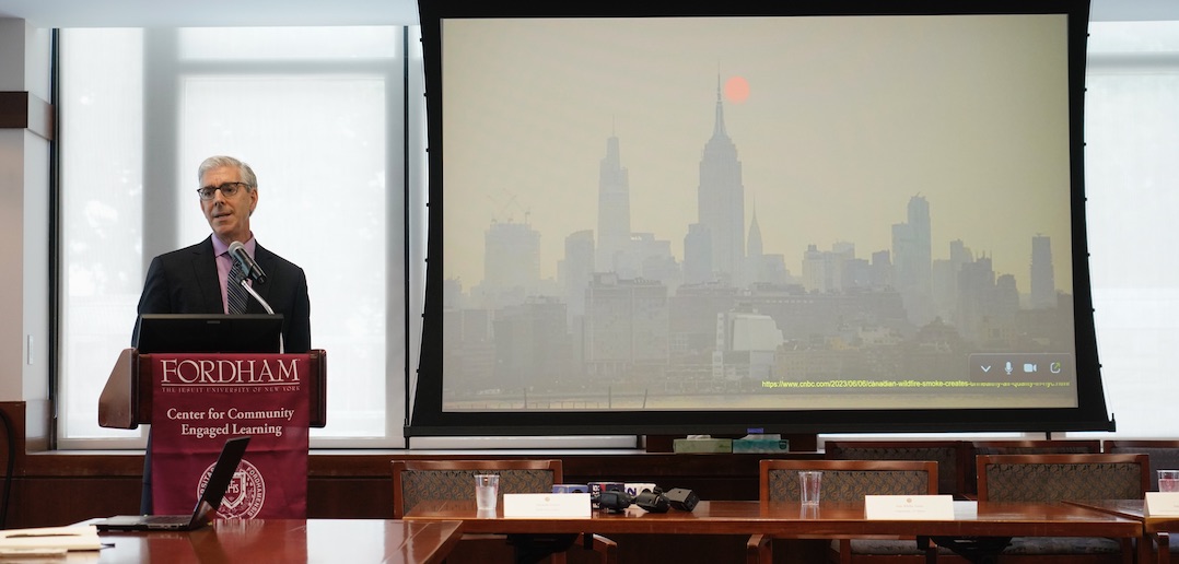 John Balbus speaks at a podium next to a presentation slide of NYC covered in smog.