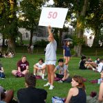 A girl holds up a sign that says "50" in the middle of a circle of students seated on the grass.