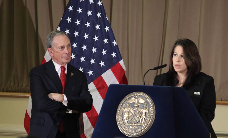 A woman in a business suit speaks at a podium with a man in a suit looking on and a United States flag in the background