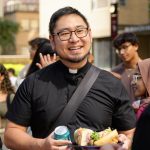 A Jesuit wearing a traditional black outfit holds food and talks with someone.