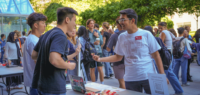 Two male presenting students engaging in conversation over club fair table.