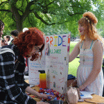 Pride Club table. Two female presenting on one side of the table, one student looking down at table.