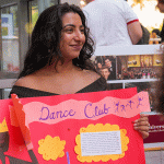 Female presenting student, smiling off into the distance holding a "Dance Club" sign.