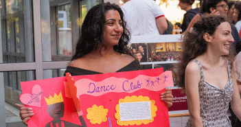 Female presenting student, smiling off into the distance holding a "Dance Club" sign.