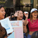 3 female presenting students, student in the middle is holding a sign. They are all smiling in mid-cheer.