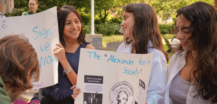 two female presenting students smiling at each other as they hold up club signs