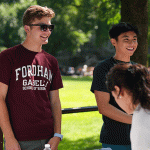 Group of students smiling. One male presenting student in Fordham shirt and sunglasses on. The other 2 beside him smiling. Greenery in background.