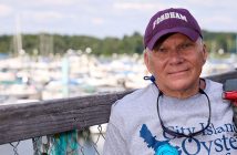 A man wearing a Fordham hat and a City Island Oyster Reef T-shirt on a dock with boats on the water in the background