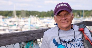 A man wearing a Fordham hat and a City Island Oyster Reef T-shirt on a dock with boats on the water in the background