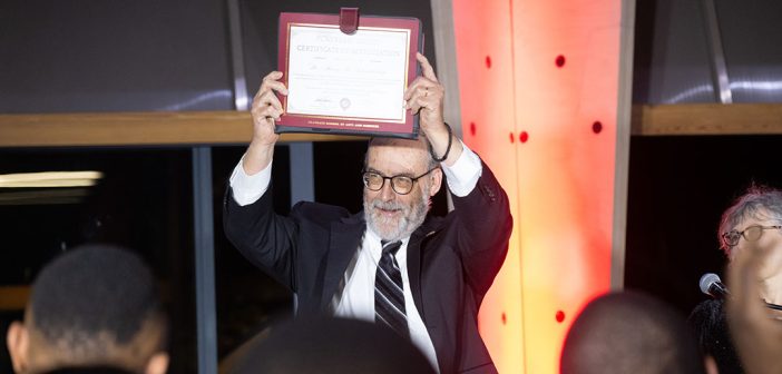 A man with a beard holds an award up over his head on stage.