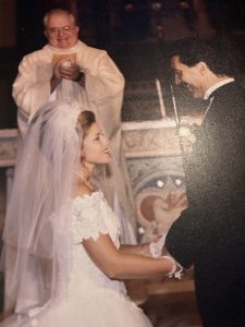 A bride and groom standing in front of a church alter, with Father Daly in the background