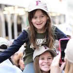 A girl wearing a Fordham baseball cap sits on the shoulders of a young woman wearing a Fordham baseball cap.
