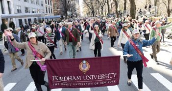 A group of parade participants hold a Fordham banner and smile and wave at the crowd.
